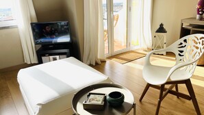 A hotel room with a bed, a television displaying a news channel, a white chair, a small table with a vase, and a view of the outdoors through a sliding door. - Boho Tavira Apartment (Tavira)
