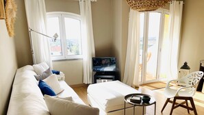 A modern hotel room with a white sofa, a small table with a tray, a TV, and a view of the outdoors through large windows. - Boho Tavira Apartment (Tavira)