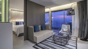 A modern hotel room with a sofa, armchair, and a round table. There is a bed with a white duvet and blue pillows. The room has a large window with a view of the cityscape at dusk. - Vip Villas Pattaya Hollywood Jomtien Beach (Pattaya)