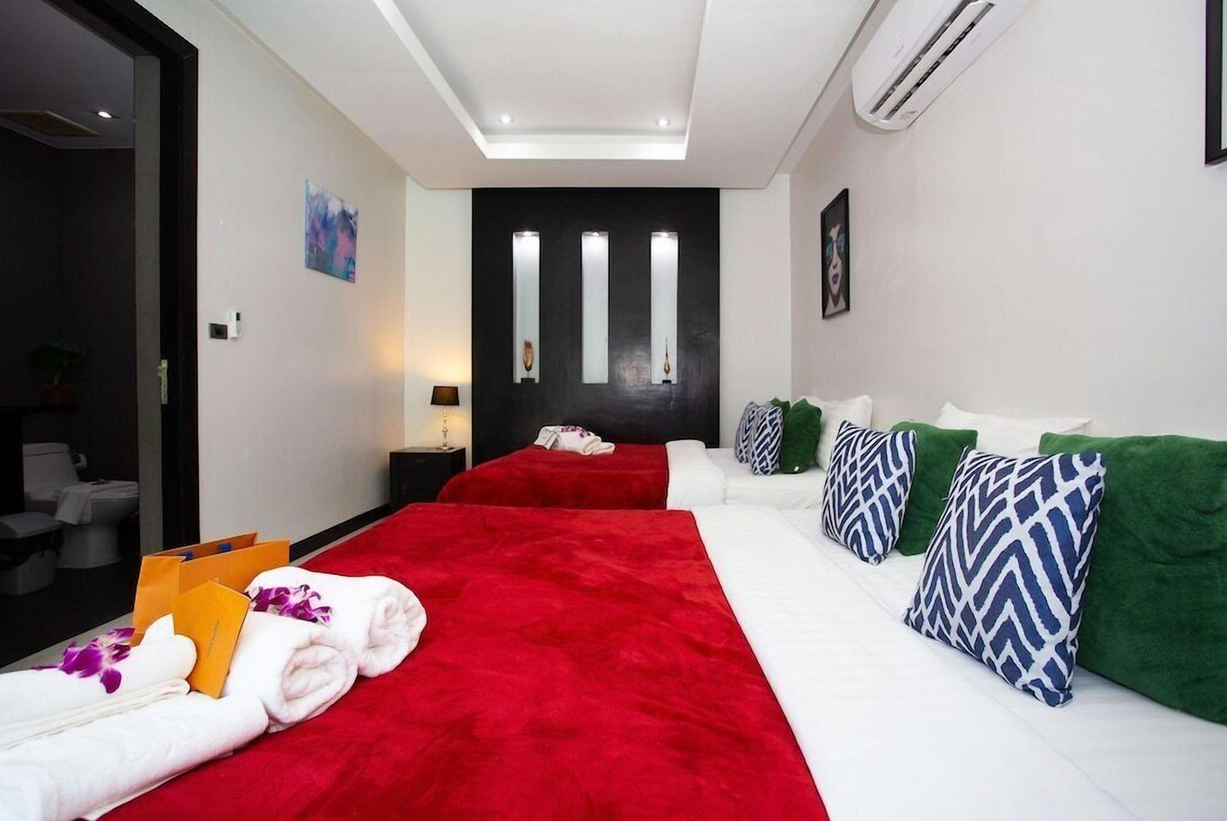 A hotel room with a red bedspread, white bedding, and green and blue patterned pillows. There is a black wall with three vertical light fixtures and a door with a dark frame.