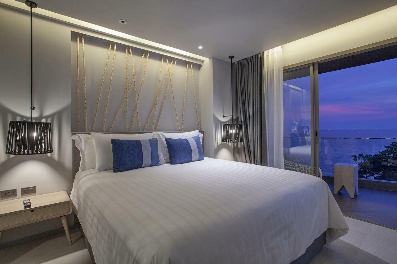 Une chambre moderne avec un grand lit, une table de chevet, une lampe et une vue sur la ville au crépuscule.