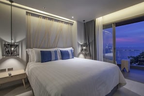 A modern bedroom with a large bed, a nightstand, a lamp, and a view of the city at dusk. - Hollywood Jomtien Beach (Pattaya)