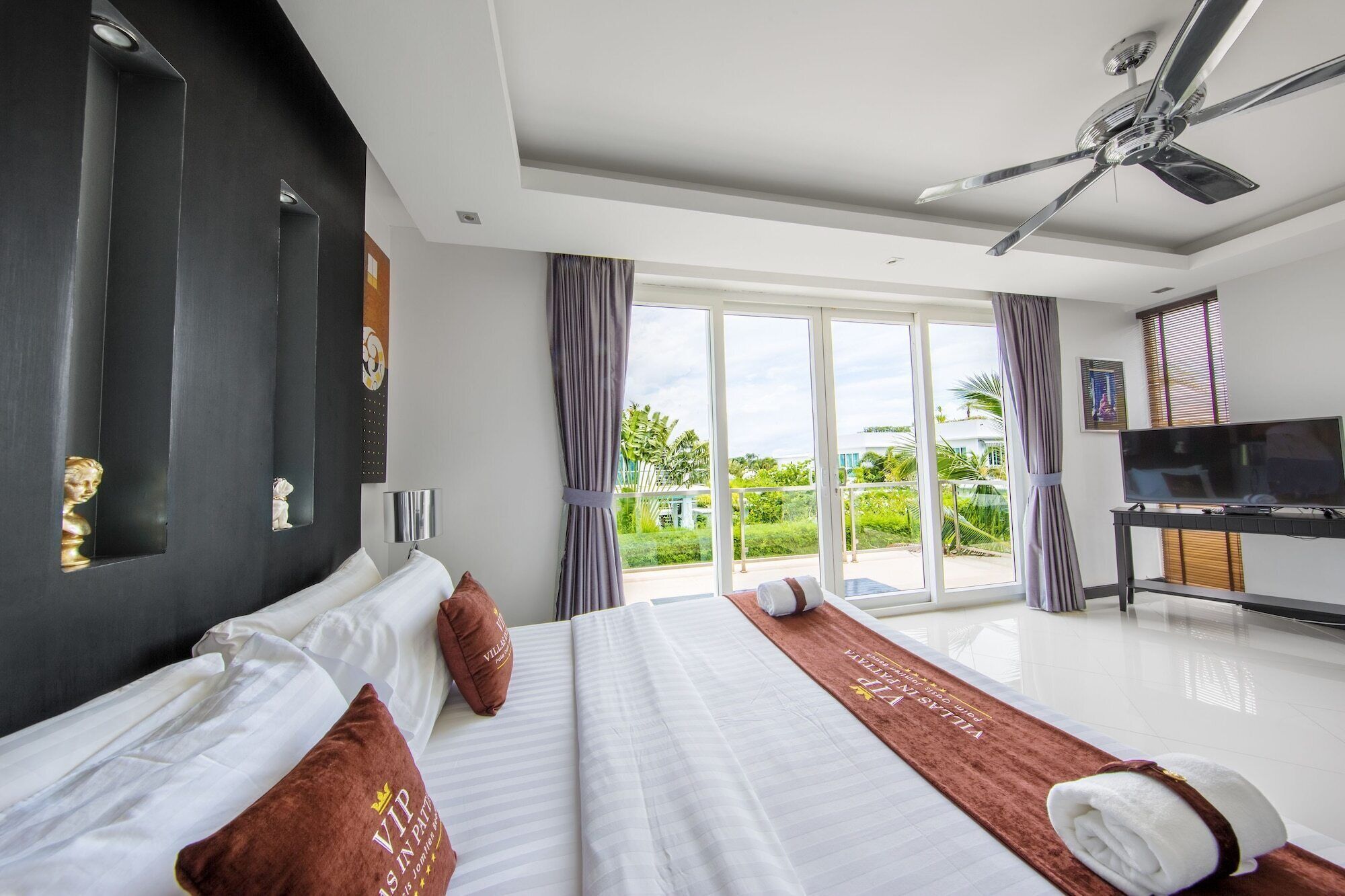 A hotel room with a large bed, a ceiling fan, a TV, and a view of greenery outside.