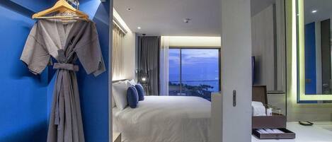 Ein modernes Hotelzimmer mit einem großen Bett, einem an der Wand hängenden Bademantel und Blick auf die Stadt im Abendrot.