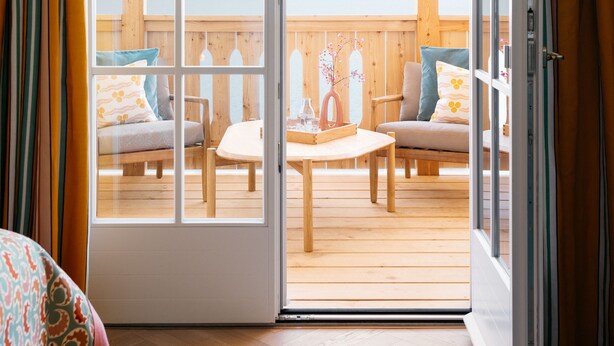 A room with a wooden floor, a bed with a patterned bedspread, and a sliding glass door leading to an outdoor deck with a wooden table and chairs. - Villa Alma - a lakeside Boutique Hotel (St. Gilgen)