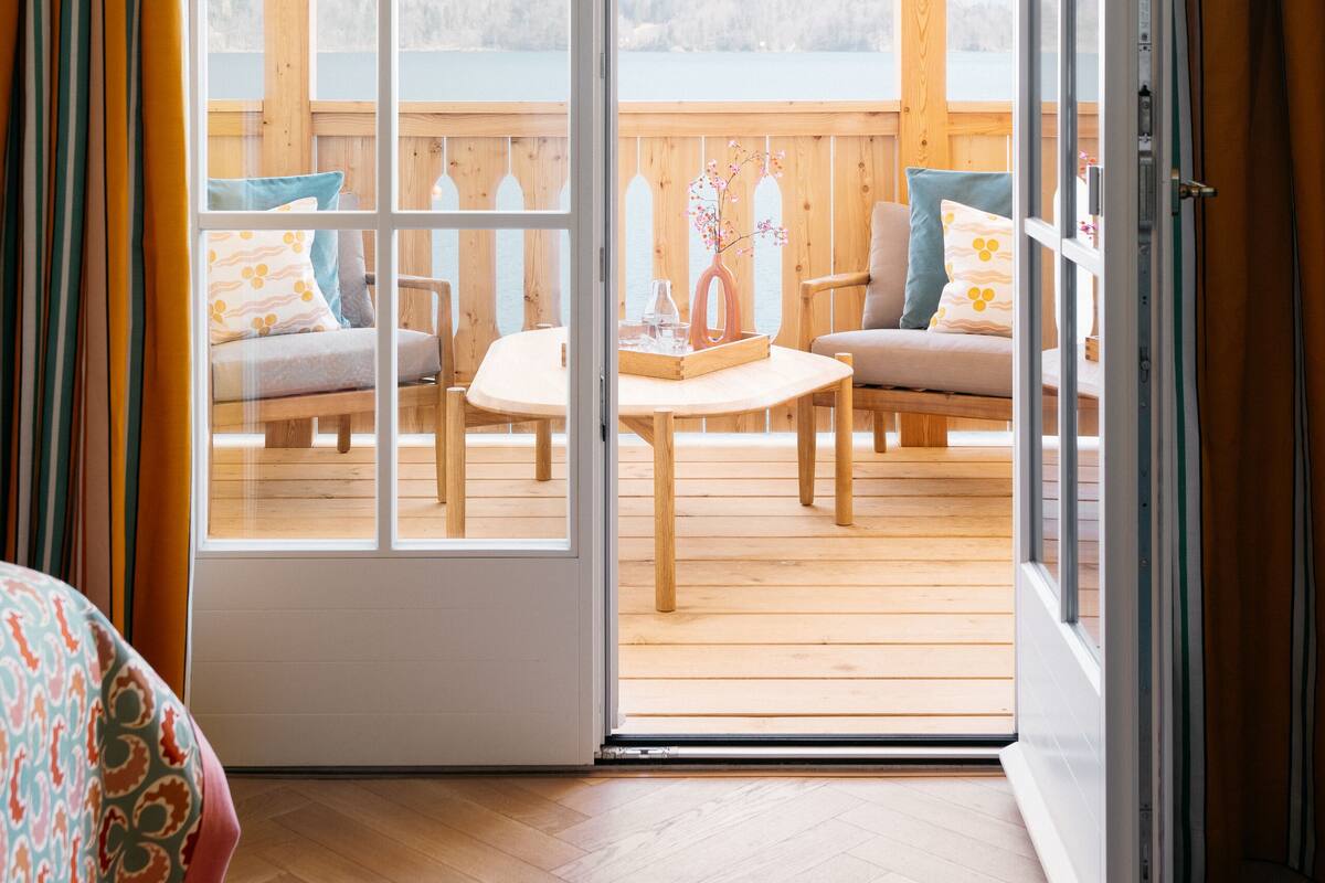 Une pièce dotée d’un plancher en bois, d’un lit recouvert d’une couvre-lit à motifs et d’une porte-fenêtre coulissante donnant sur une terrasse extérieure avec une table et des chaises en bois.