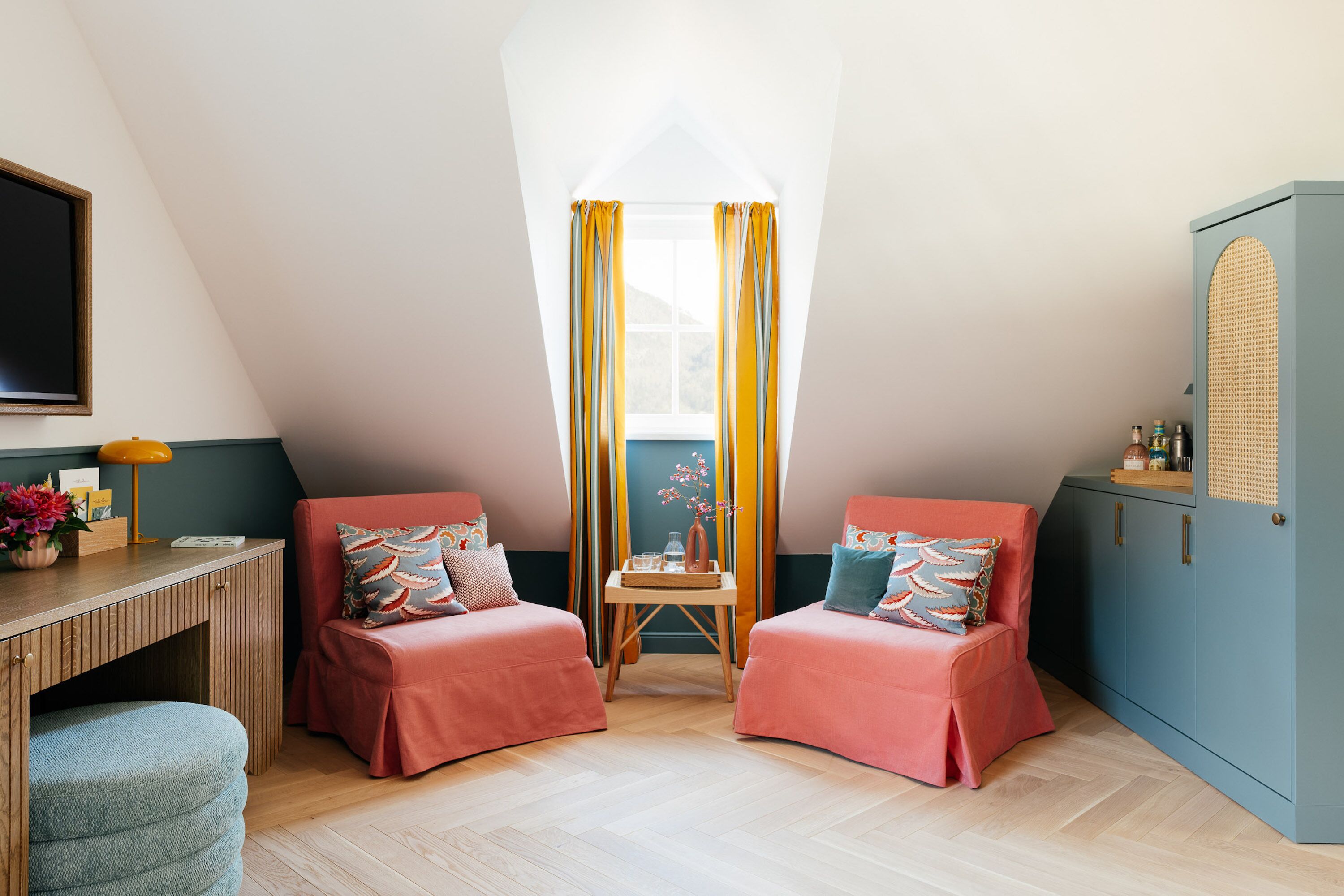 Um quarto acolhedor com duas poltronas estofadas em rosa, uma escrivaninha de madeira, uma mesinha com um vaso e um armário com porta de fibra natural.