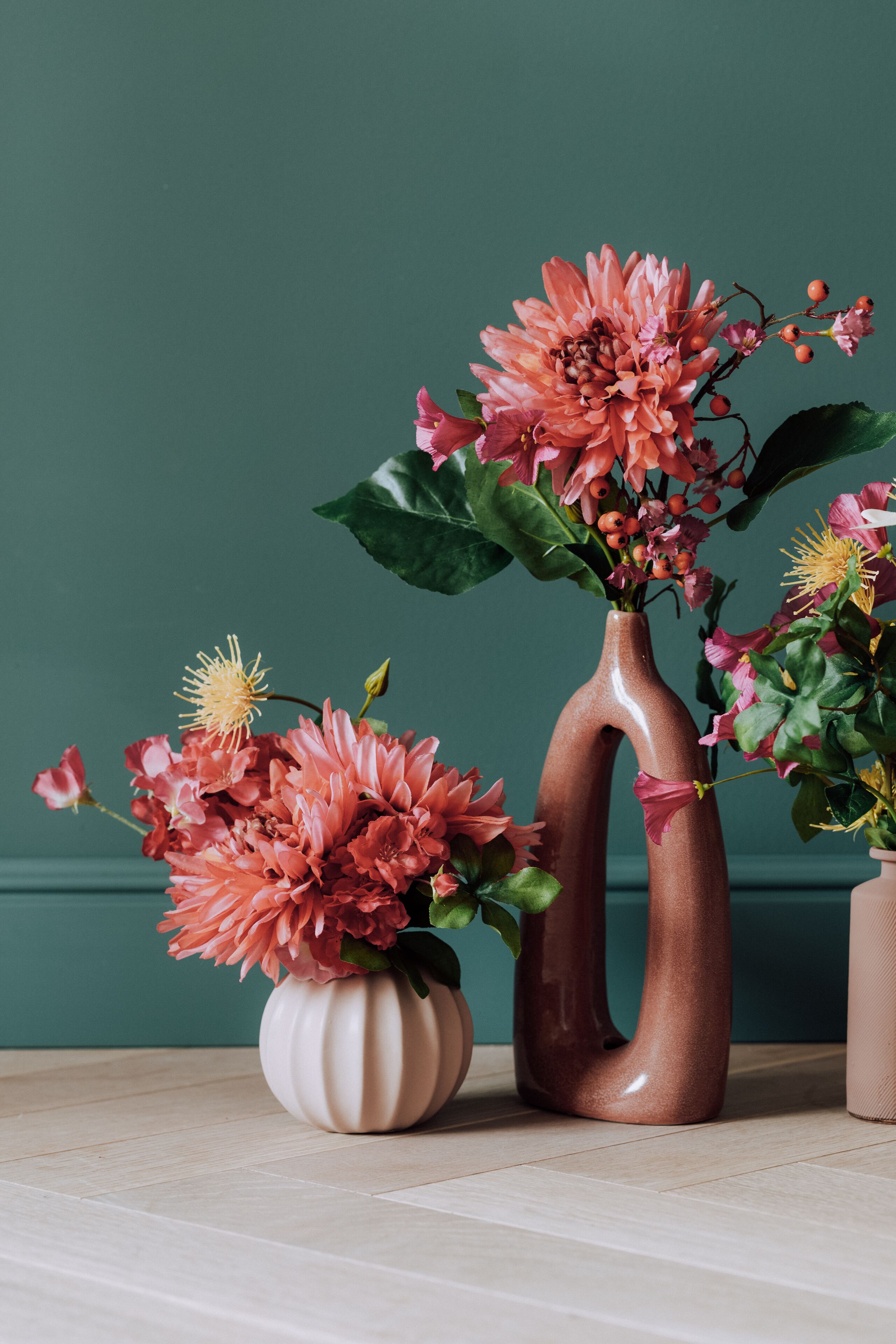 Three ceramic vases with flowers on a wooden surface against a teal wall.