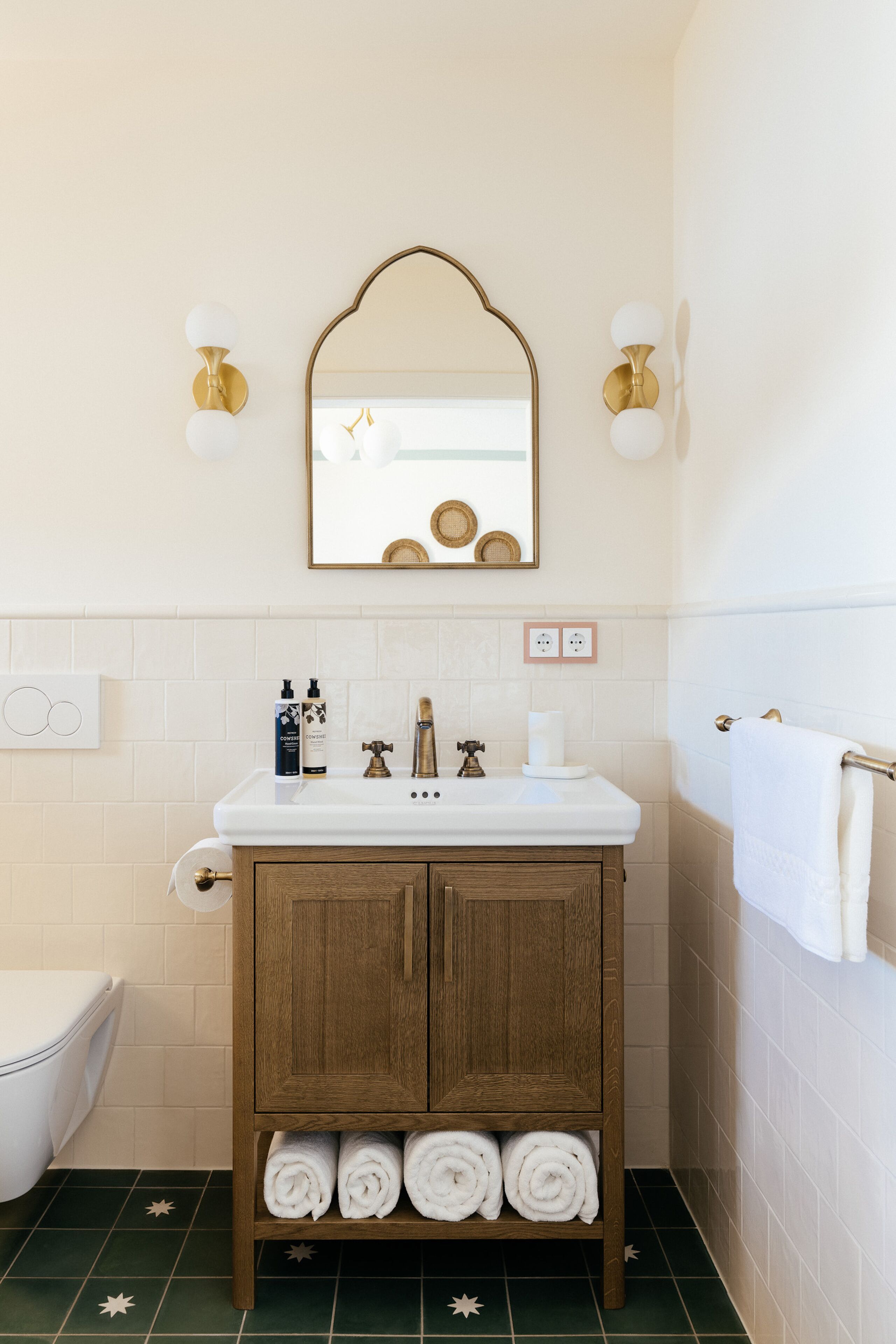 A bathroom with a white sink, wooden cabinet, and a mirror above it. There are towels rolled up in the cabinet and a toilet visible to the left.