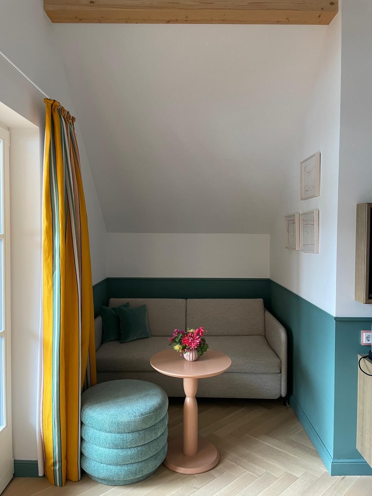 Una habitació petita i acollidora amb un sofà, una taula rodona amb un ram de flors, un otoman color turquesa i una finestra amb cortines.