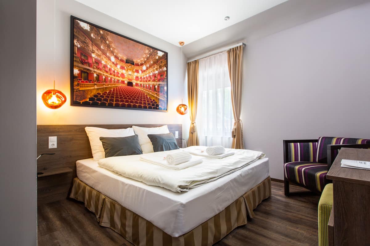 Une chambre d’hôtel avec un grand lit, une tête de lit, un coin salon avec un fauteuil à rayures et un tableau représentant un opéra, accroché au mur.