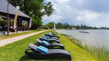 Outdoor lounge chairs with blue cushions by a lake, a path, and a building with flags.