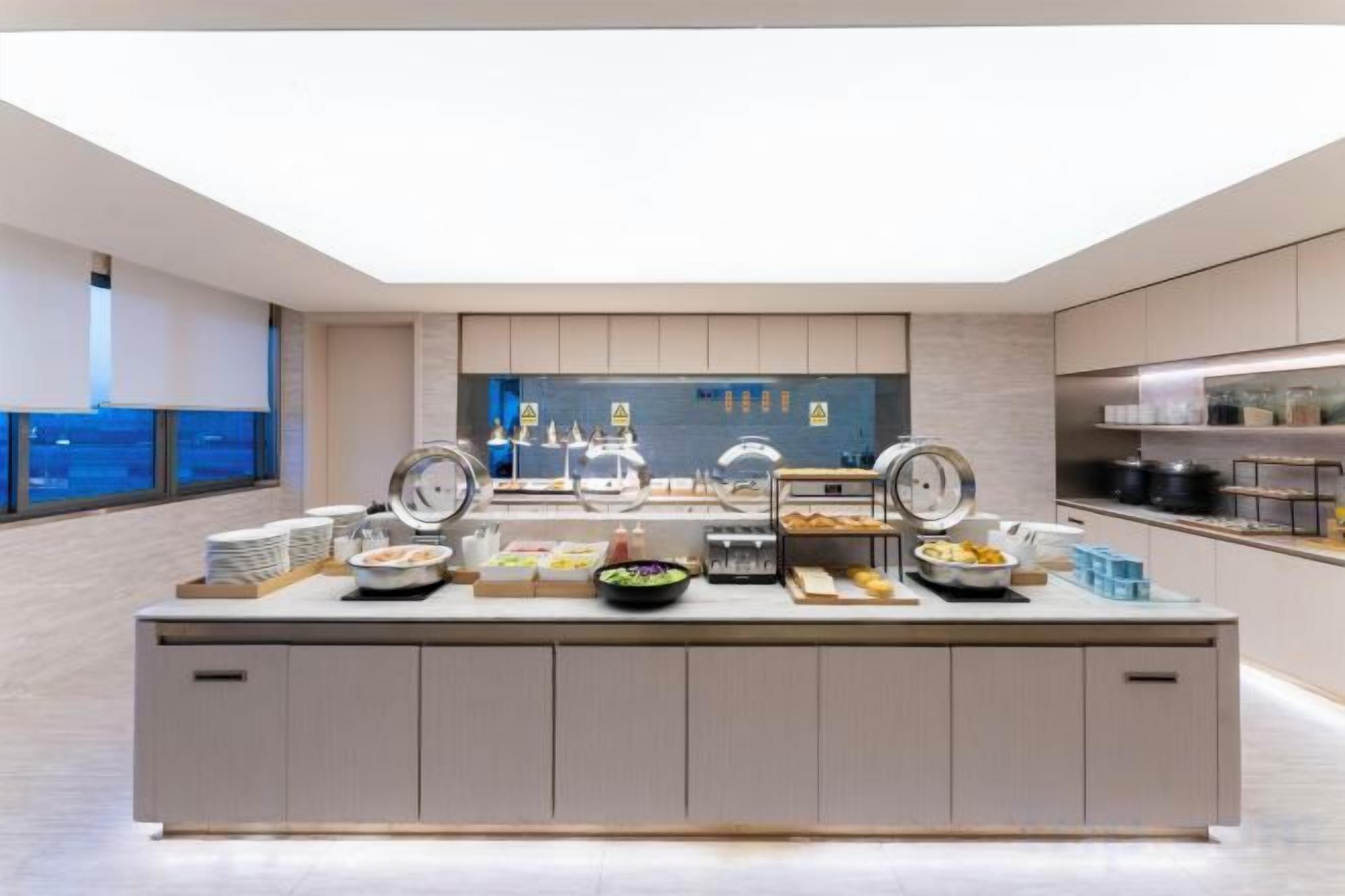 Un buffet de petit-déjeuner moderne proposant une grande variété d’aliments, avec un coin cuisine en arrière-plan et une grande fenêtre offrant une vue agréable.