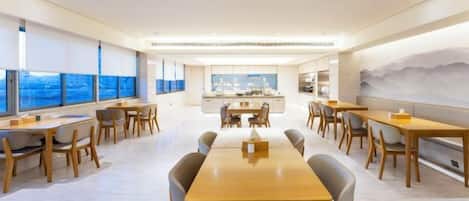 A modern dining area with a long wooden table and chairs, a kitchen area in the background, and large windows offering a view.