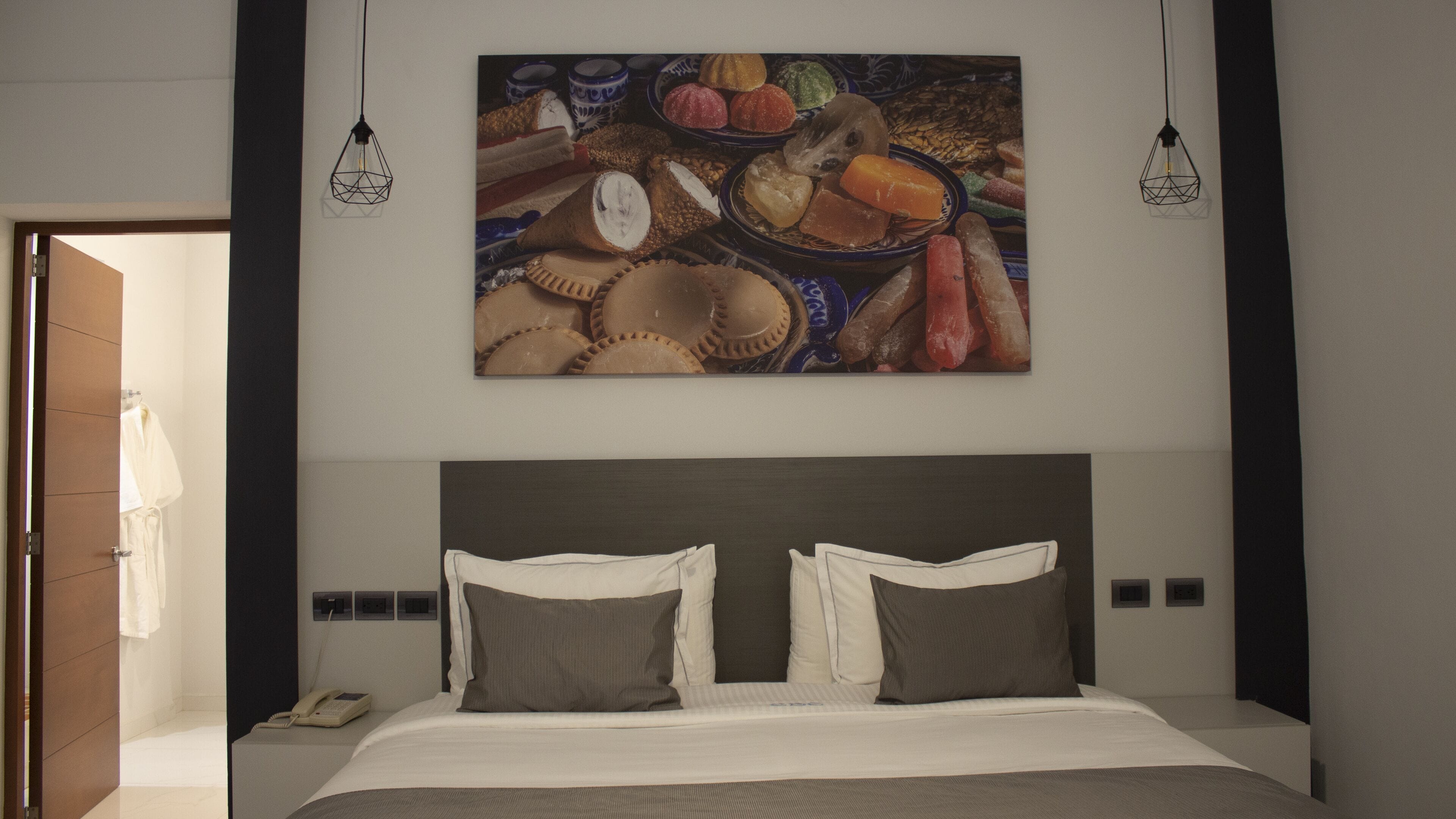 Une chambre d’hôtel moderne avec un grand lit, deux lampes de chevet et une œuvre d’art fixée au mur représentant différentes sortes de produits de boulangerie.