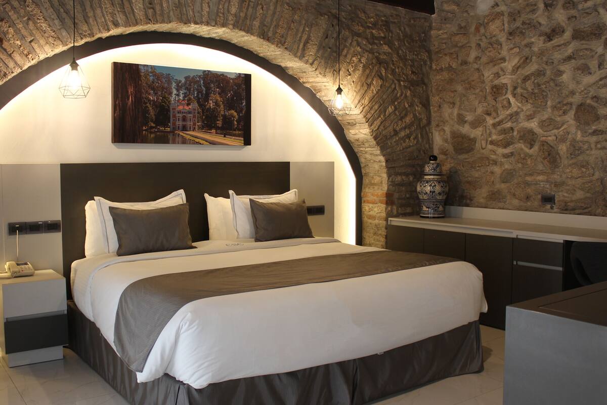 Ein modernes Schlafzimmer mit einem Steingewölbe, einem Bett mit weißer und grauer Bettwäsche, einem Nachttisch mit einem Telefon und einem Gemälde über dem Bett.