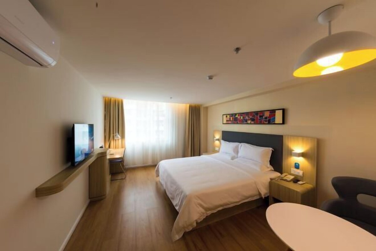 A hotel room with a bed, bedside table, lamp, TV, and a painting on the wall.