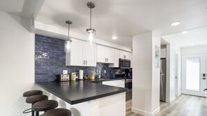 A modern kitchen with white cabinets, a black countertop, and a blue backsplash. There are three bar stools and a stainless steel refrigerator. - Sophia (Scottsdale)