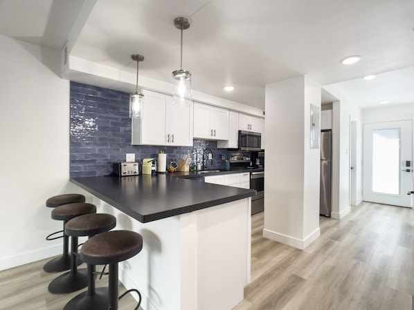 A modern kitchen with white cabinets, a black countertop, and a blue backsplash. There are three bar stools and a stainless steel refrigerator. - Sophia (Scottsdale)