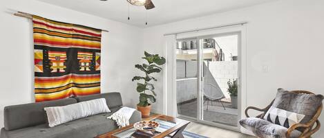 A living room with a gray sofa, a wooden coffee table, a wicker chair, a ceiling fan, and a large window with a view of a building.