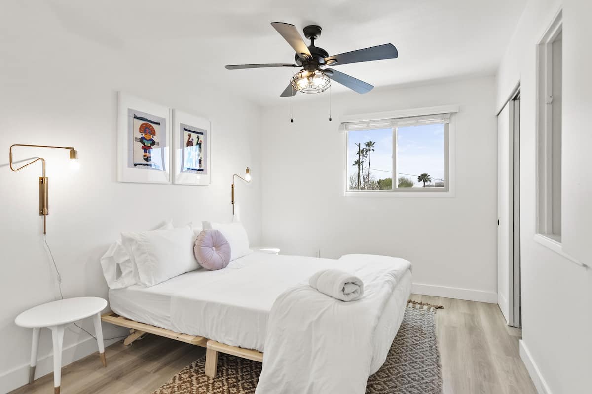 Un dormitorio con cama, ventilador de techo, ventana y una obra de arte en la pared.