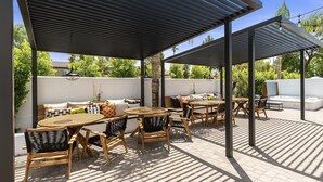 An outdoor patio area with wooden tables and chairs, a sofa, and a pergola providing shade. - Sophia (Scottsdale)