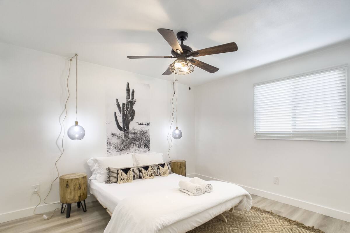Un dormitorio con cama, ventilador de techo, obra de arte de un cactus y una ventana con persianas.