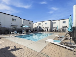 Ein Pool mit Liegestühlen und Sonnenschirmen, umgeben von einer Pflasterterrasse und einem Zaun.