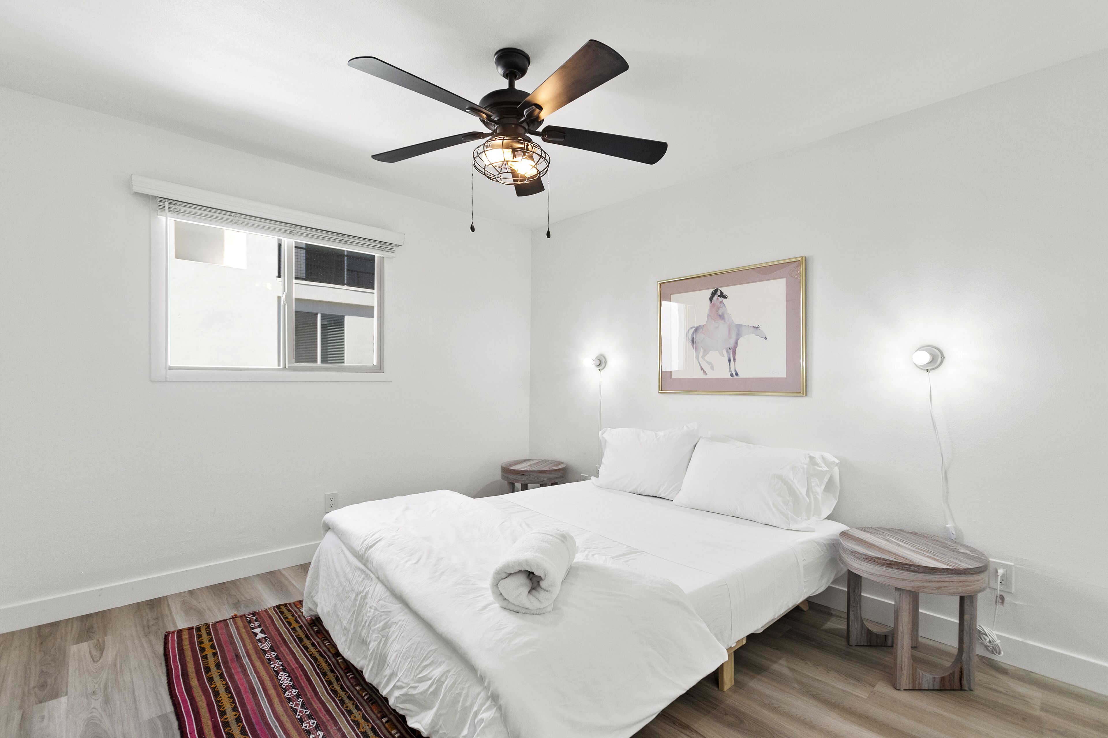 Una camera da letto con un letto, un ventilatore a soffitto, una finestra, un comodino e una lampada. Si vede anche un quadro appeso al muro.