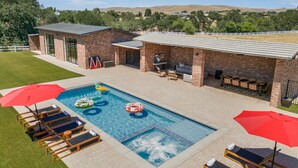 A swimming pool with lounge chairs and umbrellas, a barbecue area, and a brick building in the background. - 15 Acre Luxury Estate | VIP Experience (Paso Robles)