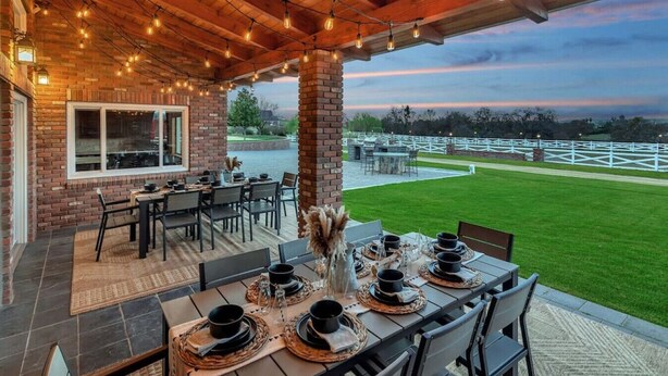 An outdoor dining area with brick walls, wooden ceiling, and string lights. Tables are set with black tableware and a vase of dried flowers. A view of a white fence and green lawn is visible in the background. - 15 Acre Luxury Estate | VIP Experience (Paso Robles)