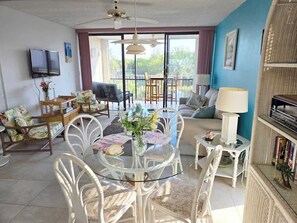 A spacious living room with a glass dining table, white wicker furniture, a large window with a view, and a flat-screen TV mounted on the wall.