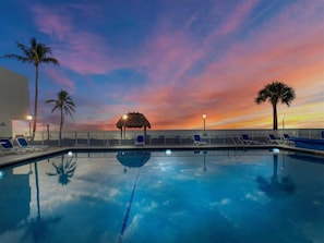 A swimming pool with lounge chairs and a thatched-roof structure at sunset.