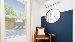 Quarto com janela, ar-condicionado, uma cadeira com almofada, espelho redondo e uma mesa pequena.