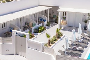 A modern outdoor seating area with lounge chairs and umbrellas, surrounded by white walls and plants. - Ambience Suites (Santorini)