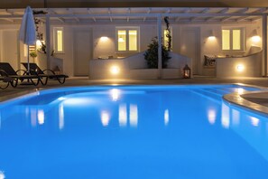 A swimming pool with a clear blue water surface, surrounded by lounge chairs and a pergola with lights. - Ambience Suites (Santorini)