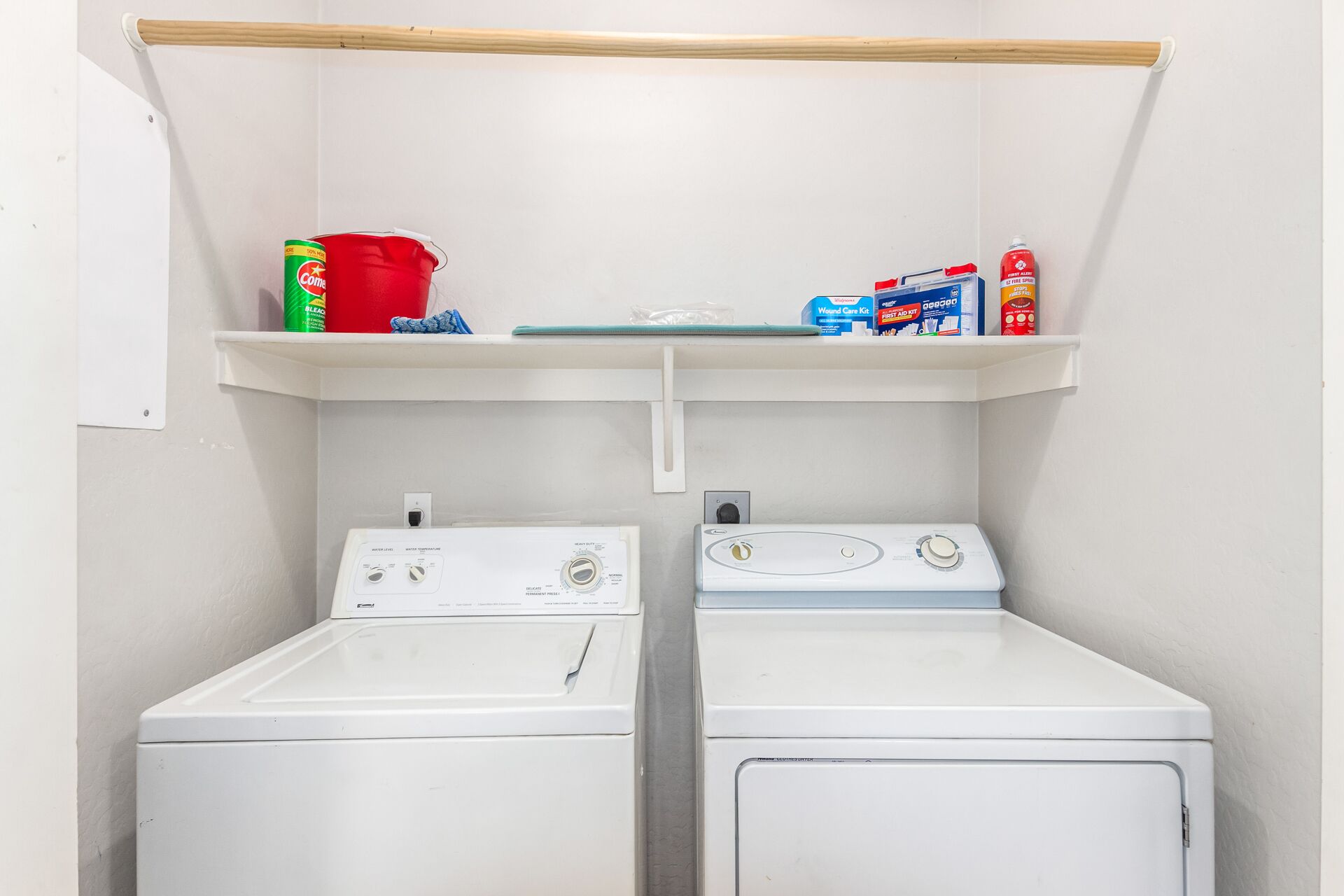 A laundry area with two white washing machines, a shelf above containing a red bucket, a green bottle, and a blue box.