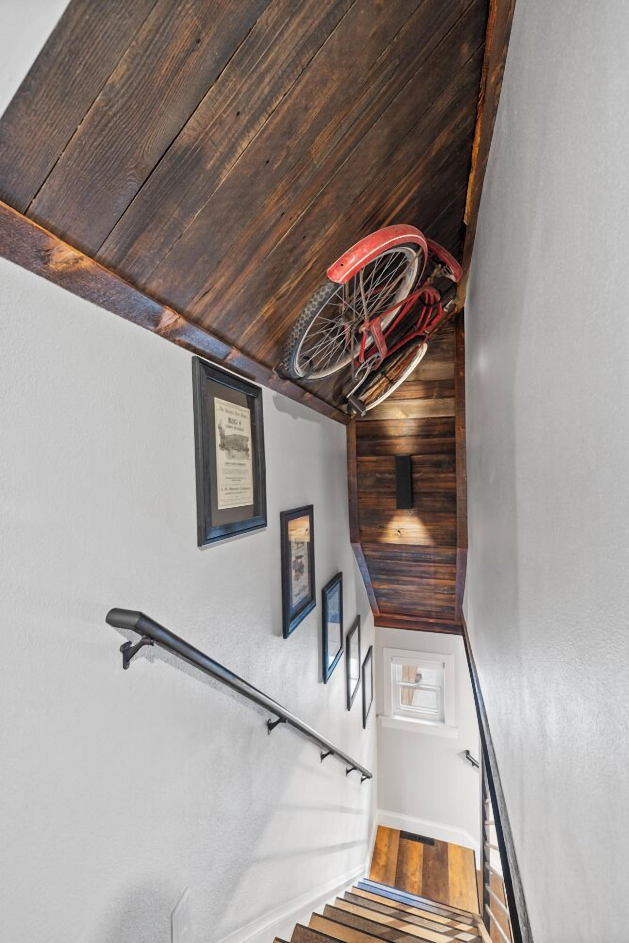 Un escalier avec une rampe en bois, un vélo rouge suspendu au plafond et des tableaux encadrés accrochés au mur.