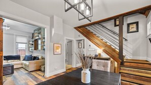 A modern living room with a dark wooden table, a white vase with dried plants, a staircase with wooden steps, and a bookshelf filled with books and decorative items. - Uniquely Modern Old Town Gem with Hot Tub (Fort Collins)