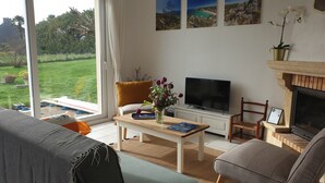 A living room with a TV, a fireplace, a sofa, a table with a vase of flowers, and a view of a garden through a large window.