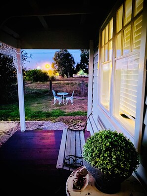 Een veranda met een bankje en een tafel, een plant in pot, en uitzicht op een grasveld met bomen en een zonsondergang op de achtergrond.
