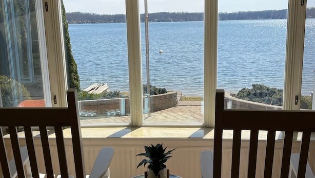 Una habitación con vistas a un lago, dos sillas de madera, una mesita con una planta y una ventana con persianas.