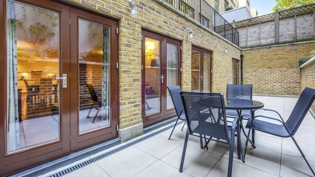 Un patio con un edificio de ladrillo, puertas de cristal grandes y una mesa con sillas.