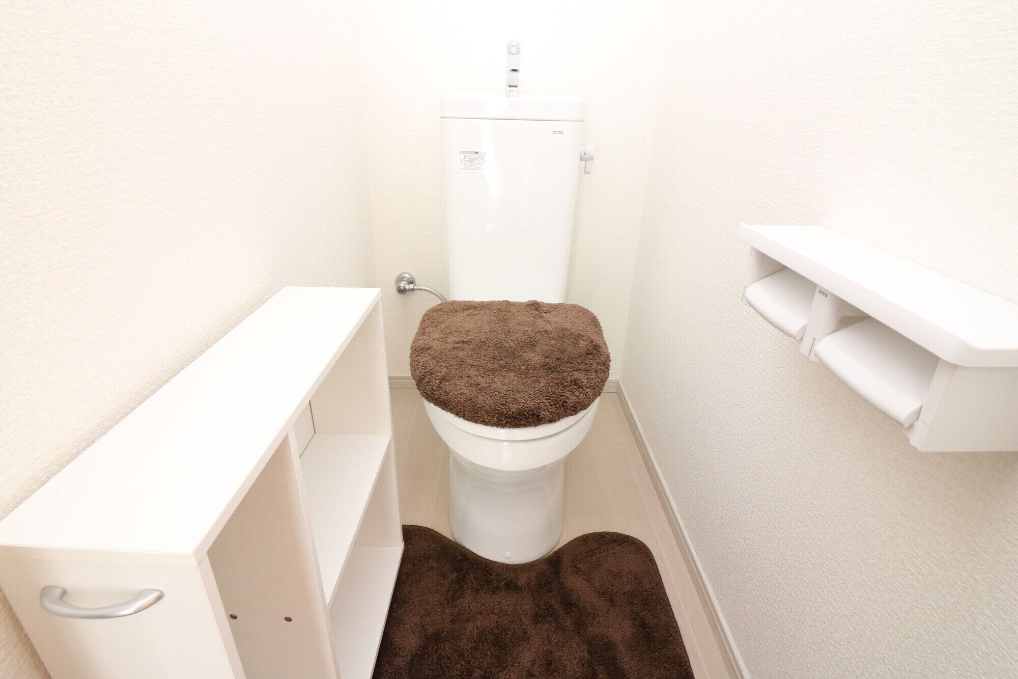 A small, white bathroom with a toilet, a white shelf, and a brown bath mat.