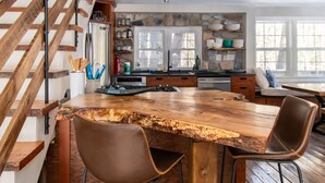 Une cuisine dotée d’un îlot central en bois rustique, d’un pare-feu en pierre et d’une cuisinière encastrée. Un escalier en bois se trouve à gauche, et une fenêtre offre une vue sur les arbres.