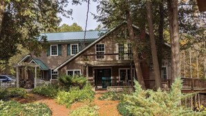 Une maison rustique en bois, dotée d’un porche couvert, entourée d’une végétation luxuriante et bordée d’un chemin en briques.