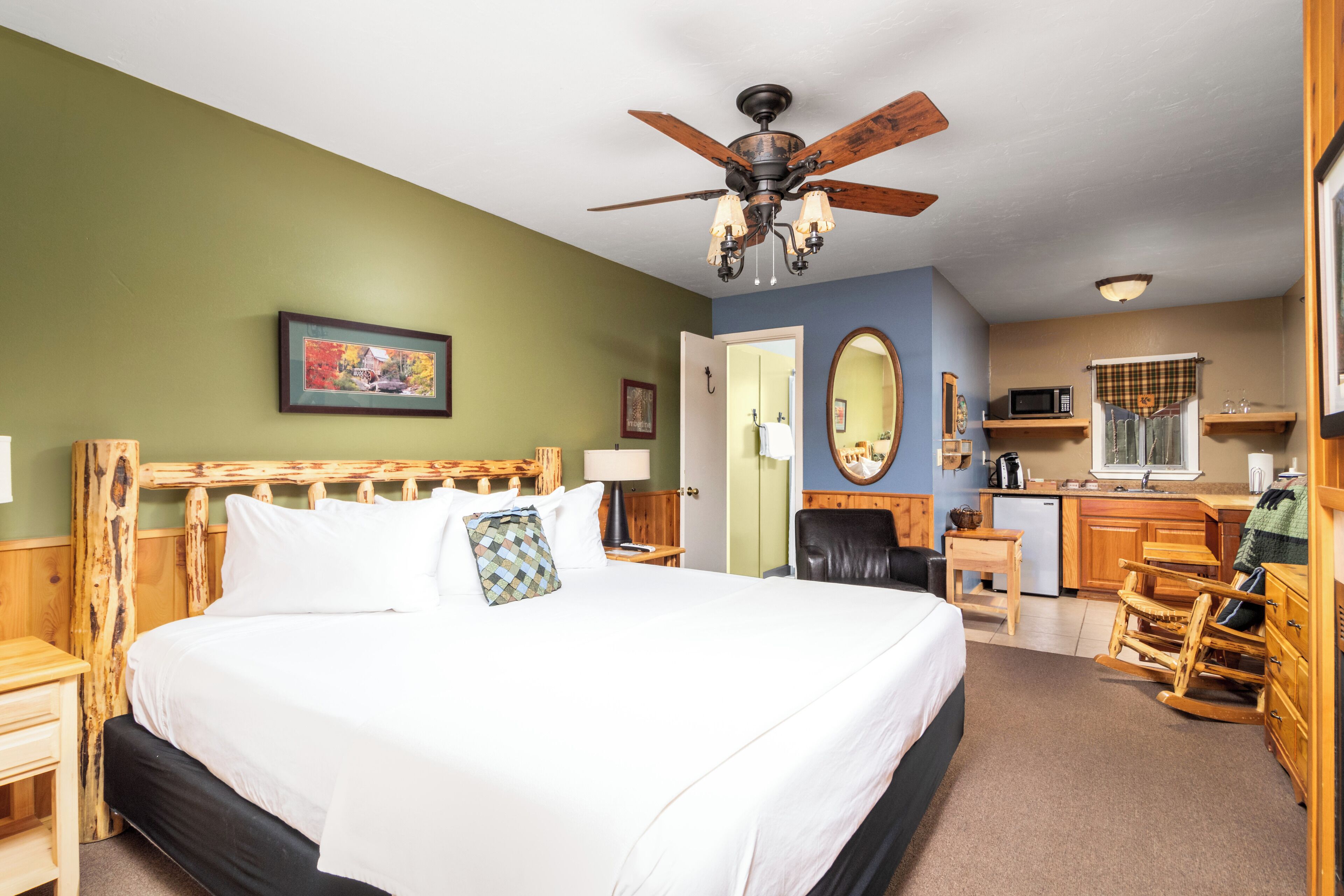 A hotel room with a large bed, a ceiling fan, a wooden headboard, a desk with a chair, a microwave, and a sink.