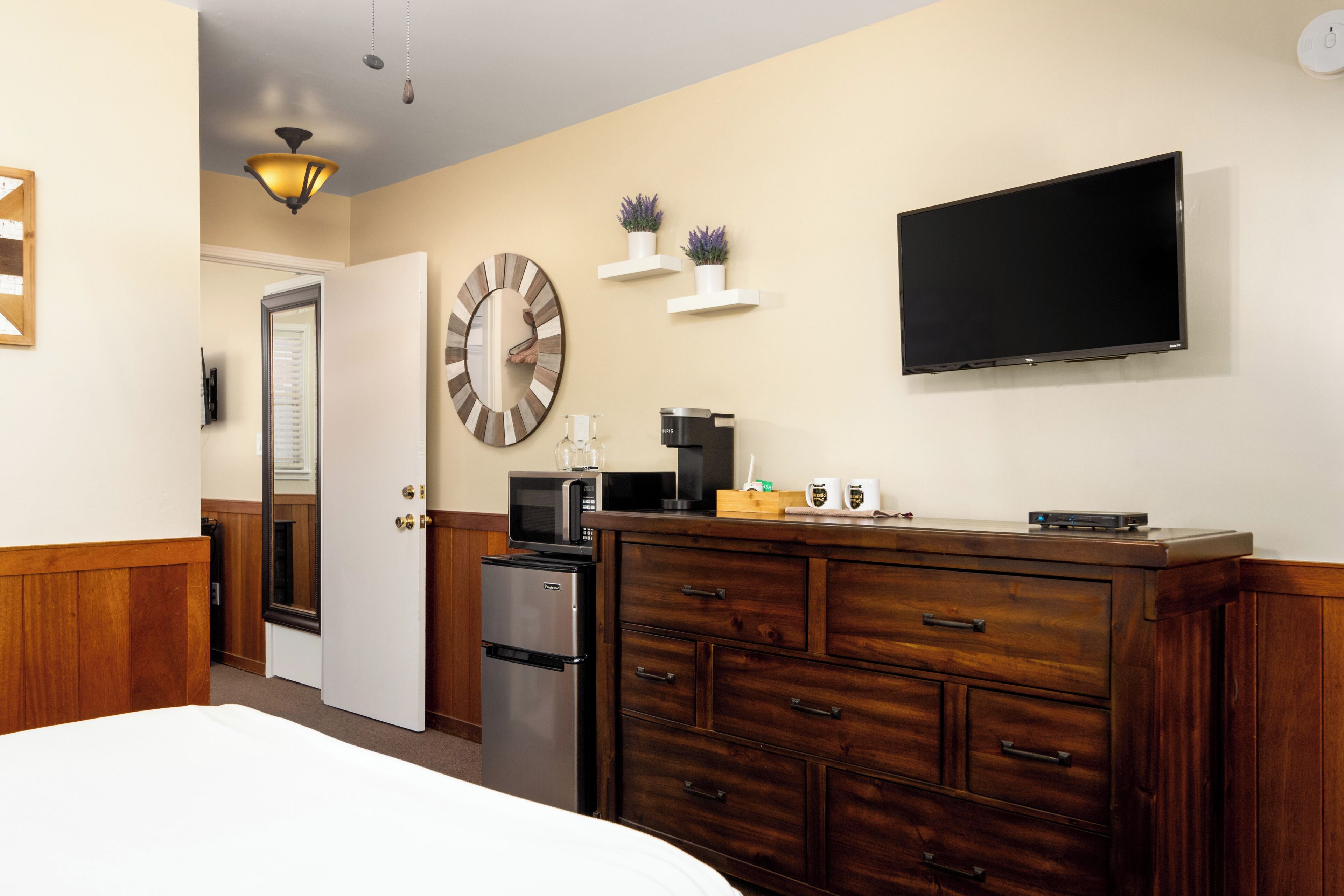 A hotel room with a bed, a wooden dresser, a microwave, a coffee maker, a flat-screen TV, and a clock on the wall.