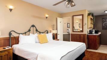Quarto de hotel com uma cama grande, um ventilador de teto, uma mesa de cabeceira de madeira com luminária, cômoda e espelho.