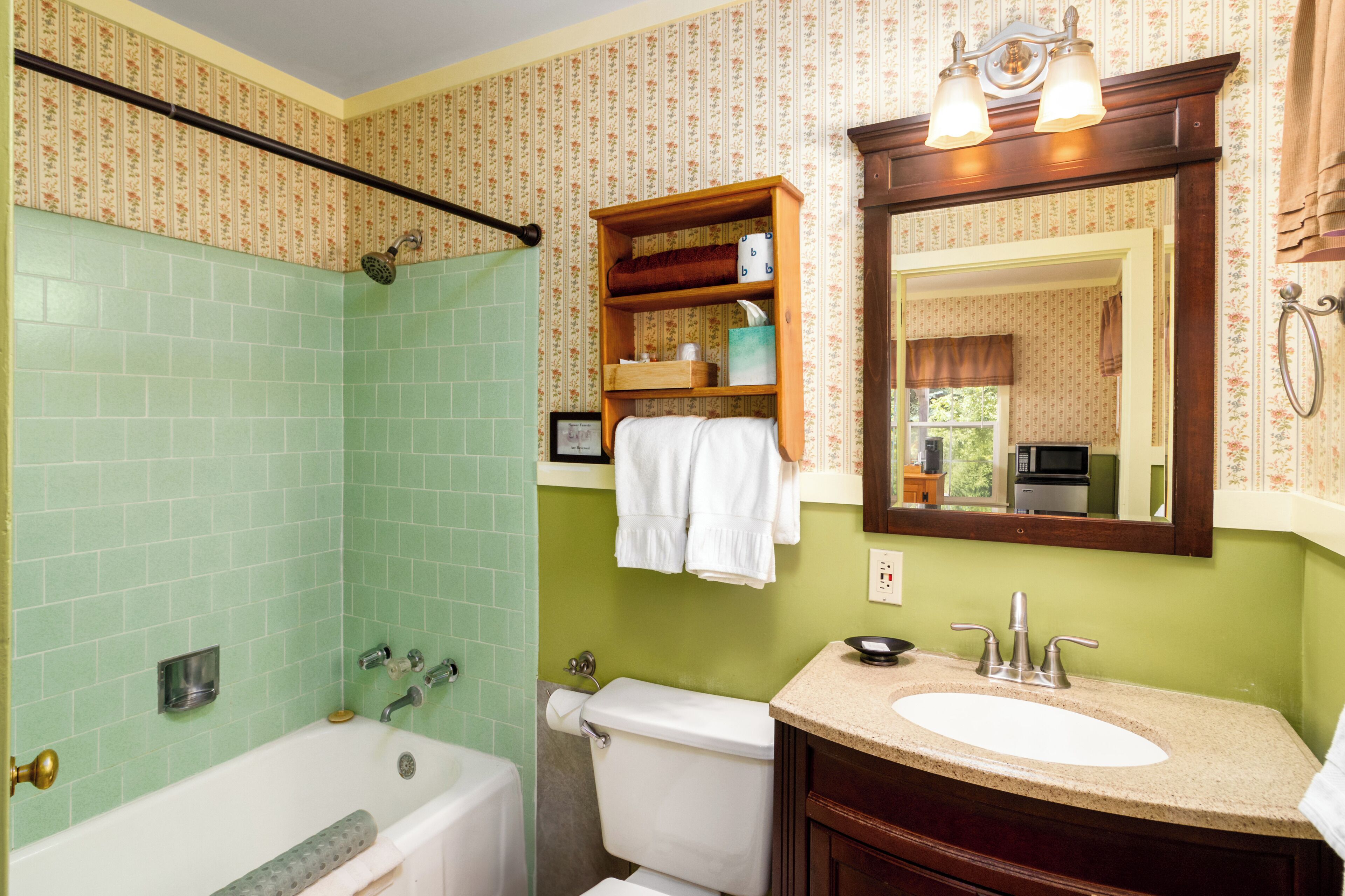 A bathroom with a green tiled shower, a white toilet, a sink with a mirror above it, and a wooden shelf with towels.
